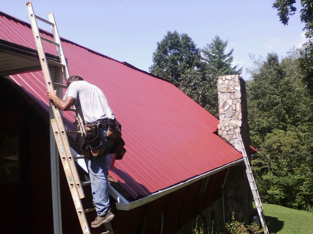 Roofer on a ladder beside a red metal roof and stone chimney — Soffit & Fascia Repair by Roofing Brothers in Marcus